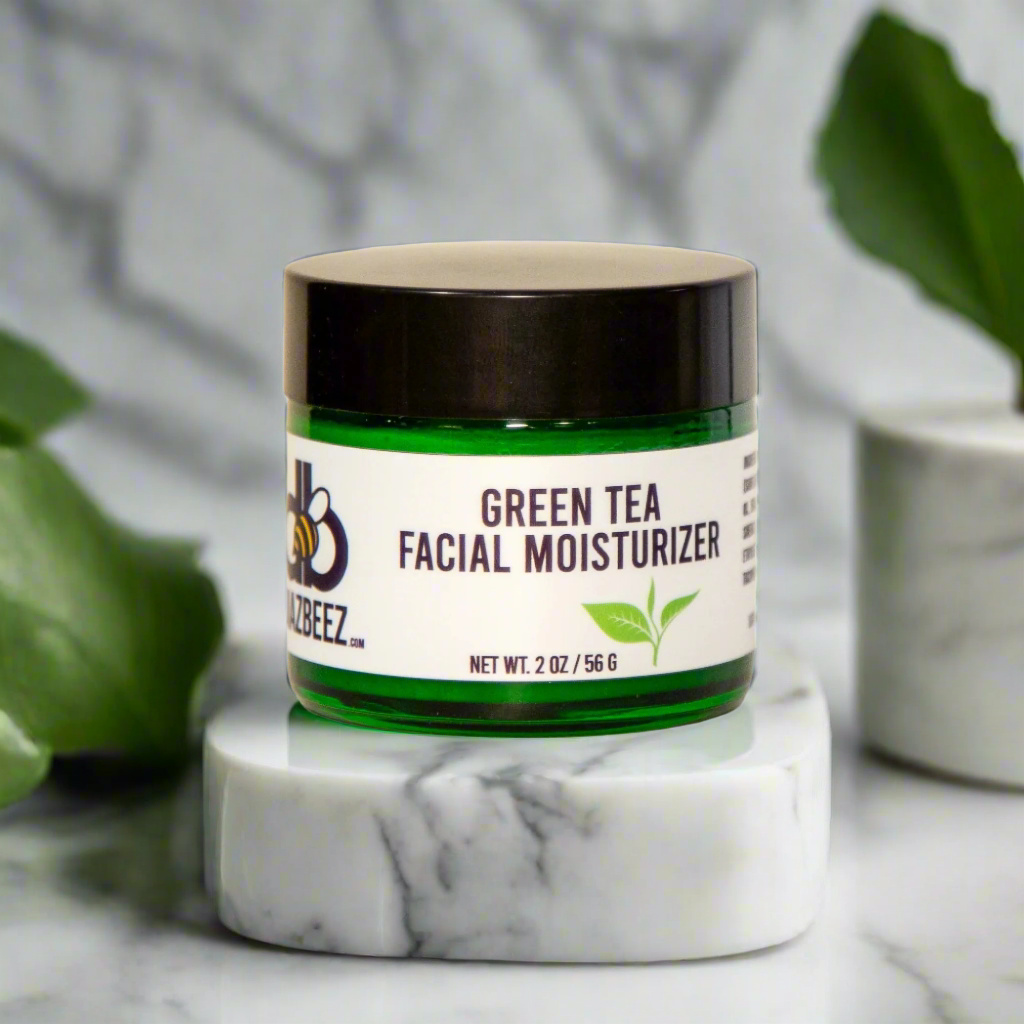 A jar of Diaz Beez Green Tea Facial Moisturizer, providing antioxidant protection and enriched with vitamin E, sits elegantly on a marble surface. The green jar with a dark brown lid rests on a small platform, surrounded by decorative green leaves.