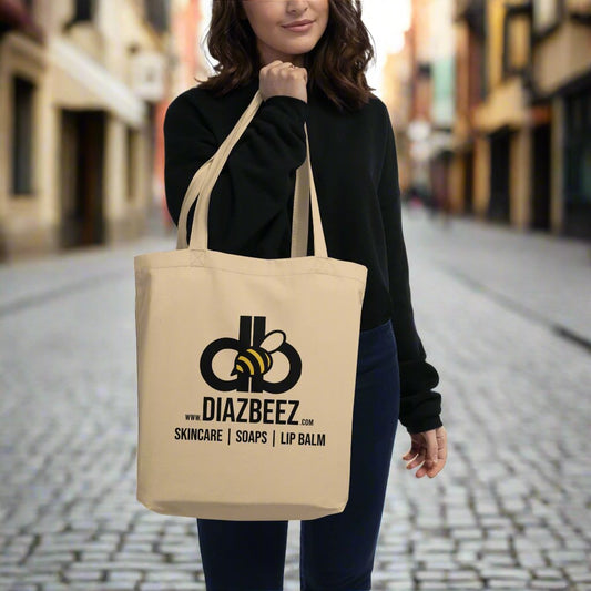 A woman in a black sweater holds an eco-friendly tote by Diaz Beez, featuring a bee illustration. The beige bag, made from organic cotton, showcases the text: "Skincare | Soaps | Lip Balm.